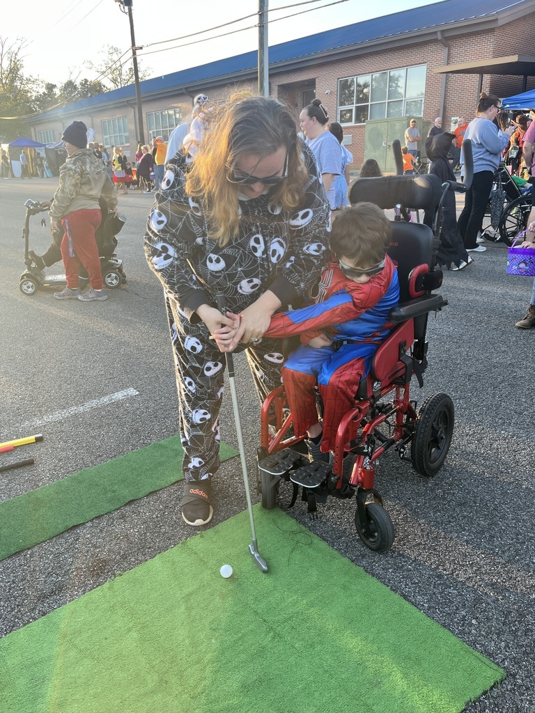An adult helps a student in a spider man costume hold a golf club while he plays put-put. The two stand in a crowded parking lot where a halloween party is being held.