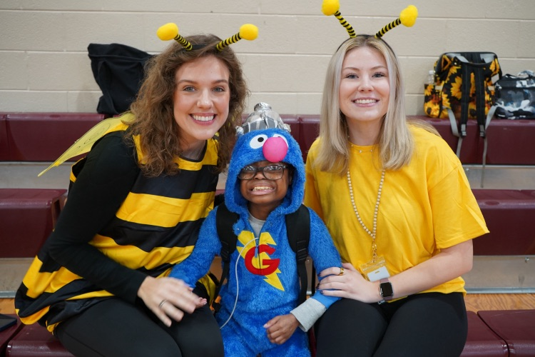 Two women in bumble bee costumes pose with a child in a Super Grover costume in a school gym.
