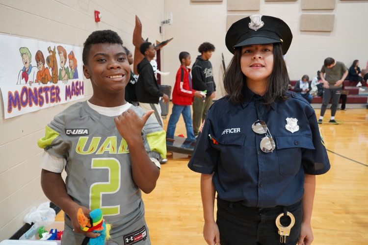 Two students dressed in costumes stand for a photo in a school gym. One student is wearing a football jersey and the other student is wearing a police officer costume  