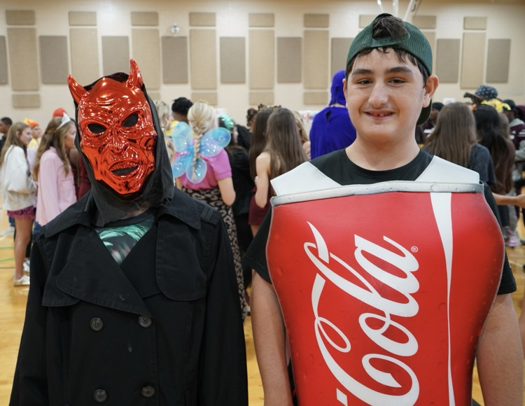 Two students stand together for a photo in a school gym. One student is wearing a scary Halloween mask. The other student is dressed like a can of Coca-Cola  