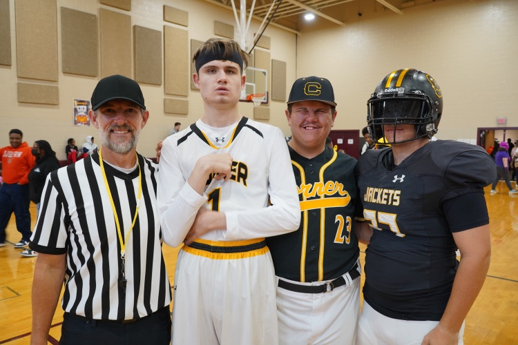 An adult wearing a referee costume poses for a photo with students wearing sports uniforms. One student is dressed like a basketball player, one is dressed like a baseball player and another student is dressed as a football player.