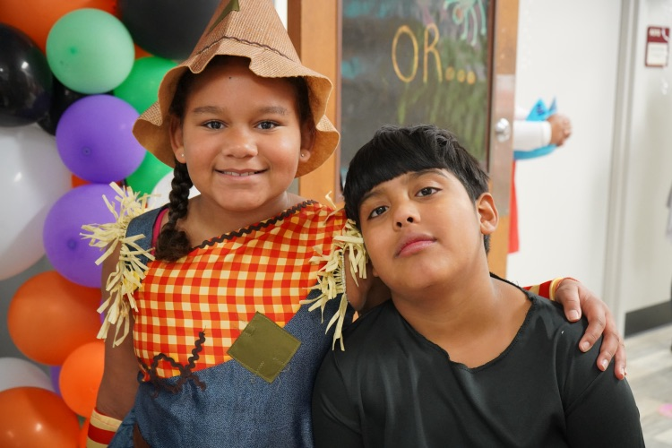 A girl and a boy stand together and smile. The girl is in a scarecrow costume