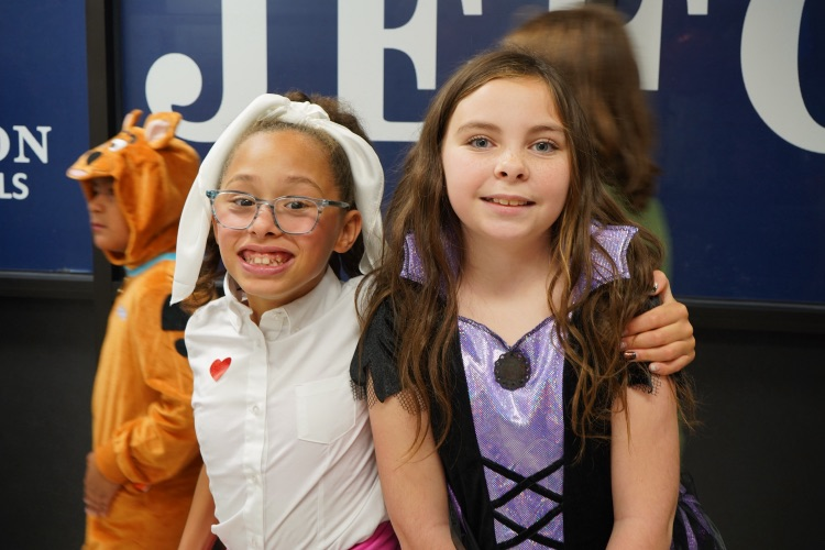 Two girls wearing Halloween costumes stand together in a hallway and smile.