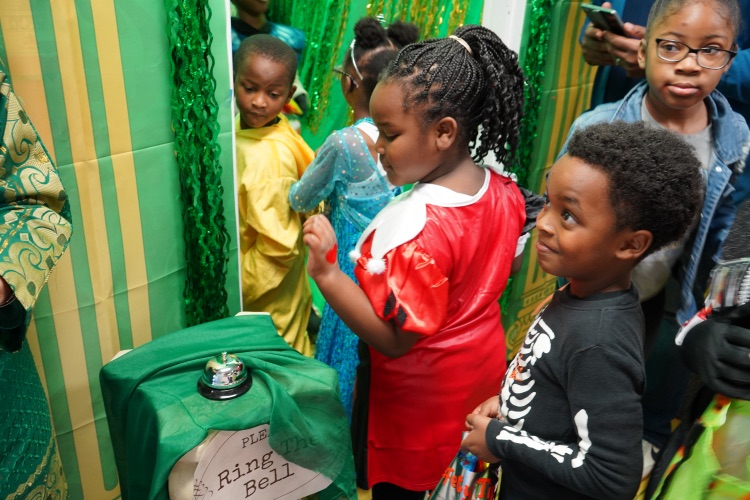 Students walk into a room decorated in green. As they walk in, some of the students ring a bell