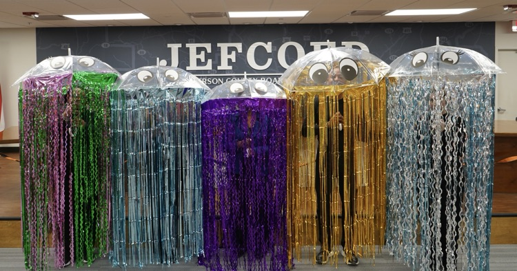 A group of board employees stand together for a photo. They’re wearing Halloween costumes made out of umbrellas and streamers to look like jellyfish. 