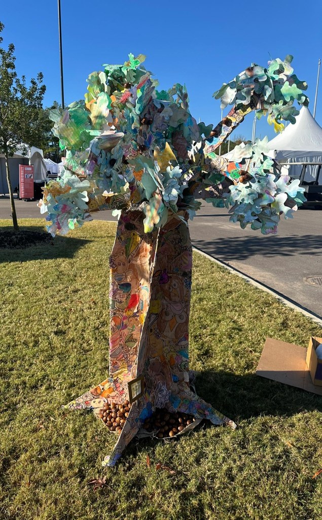 A tree sculpture made out of student art work sits on grass at a festival.