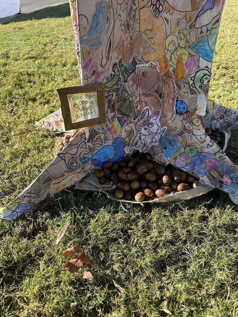 The image is a close up shot of a tree sculpture made out of student artwork at a festival. On the sculpture, there's a sign that says "Please take one Adopt a Nut" next to free nuts.