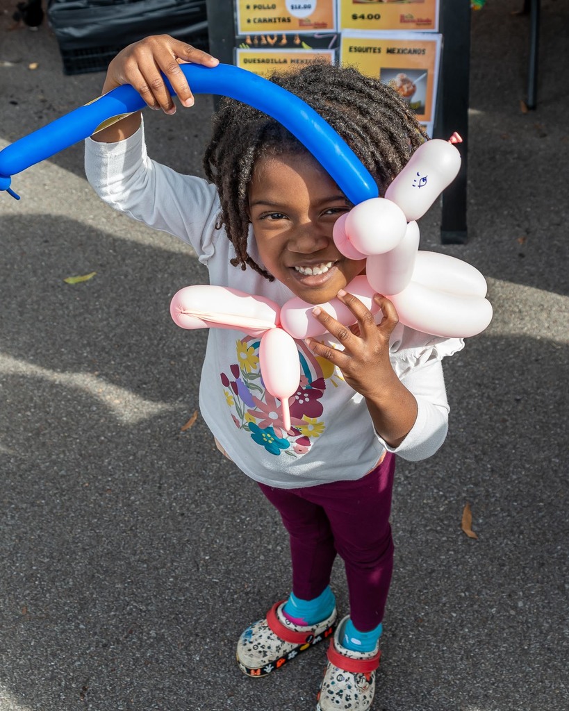 A little girl smiles while holding a balloon animal shaped like a dog on a leash while standing in a school parking lot.