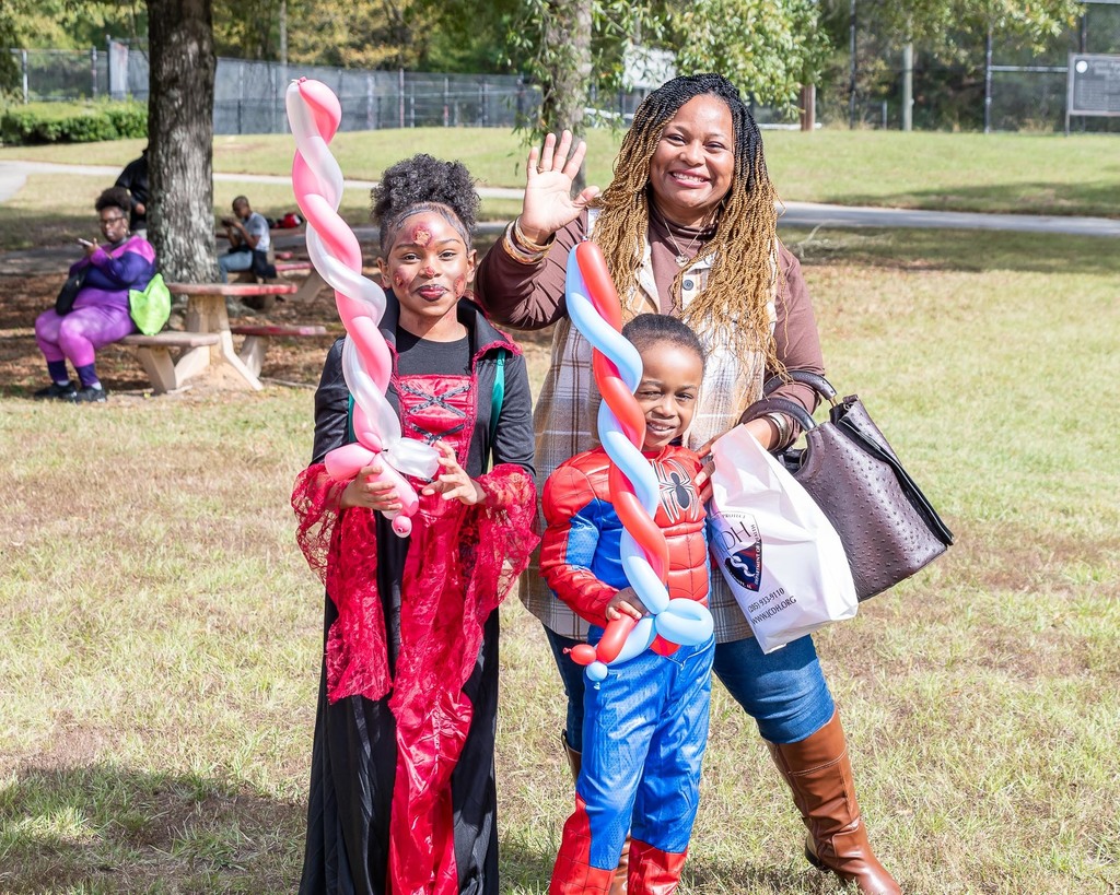 An adult and two children pose for a photo in a park. The children are wearing halloween costumes and holding up balloons shaped like swords.