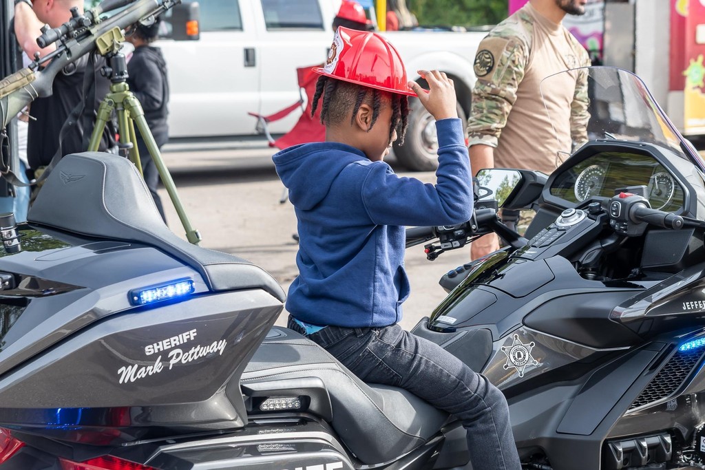 A child sits on a motorcycle while weaing a plastic firefighter hat. 