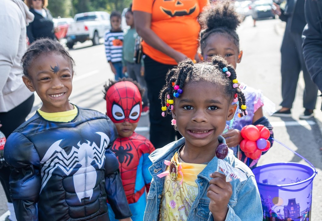 Four children pose for a photo while standing in a parking lot. The children are wearing Halloween costumes and holding buckets of candy,