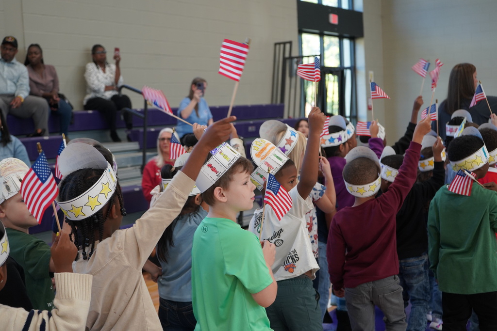 Students walk through a gym while waving small American flags and wearing matching hats that are made of paper and colored on with crayon.