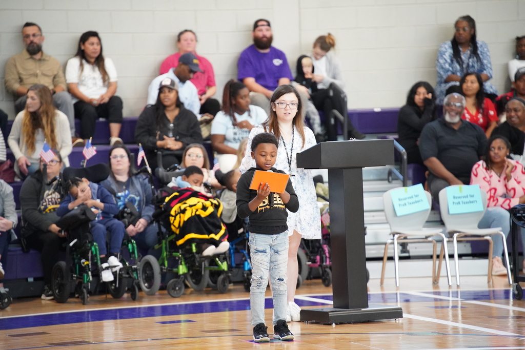 A boy reads from a piece of paper as he gives a presentation to an audience inside a school gym.