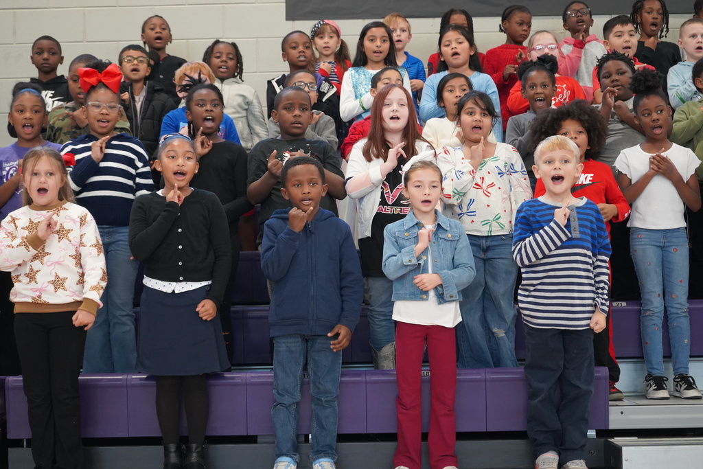 Students sing a song while standing on bleachers in a school gym.