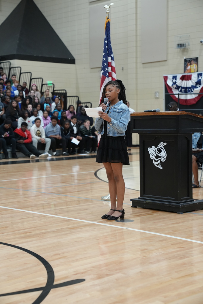 A girl speaks into a microphone while making a speech in front of a crowd of people in a school gym.