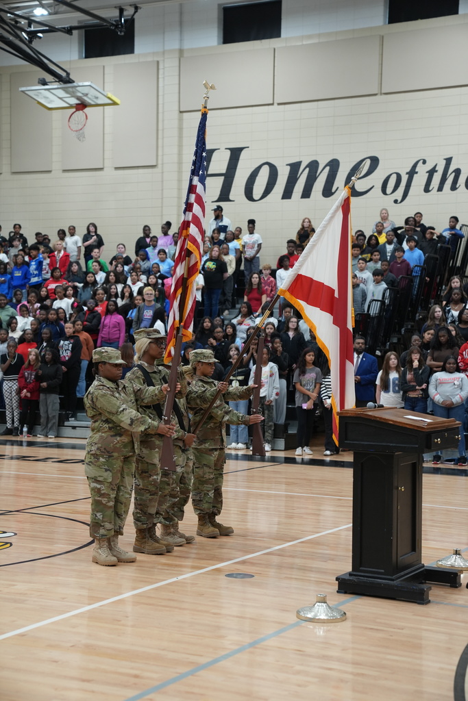 JROTC members hold up the American flag and Alabama state flag while standing in front of a crowd in a gym.