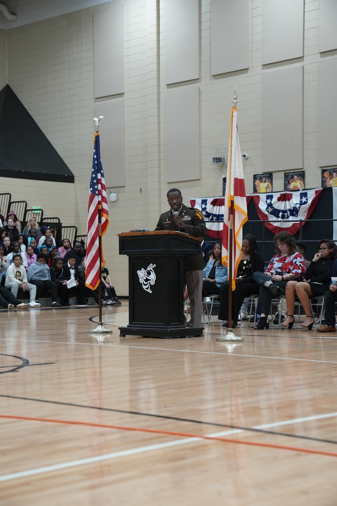A man in a military uniform speaks to a crowd in a school gym.