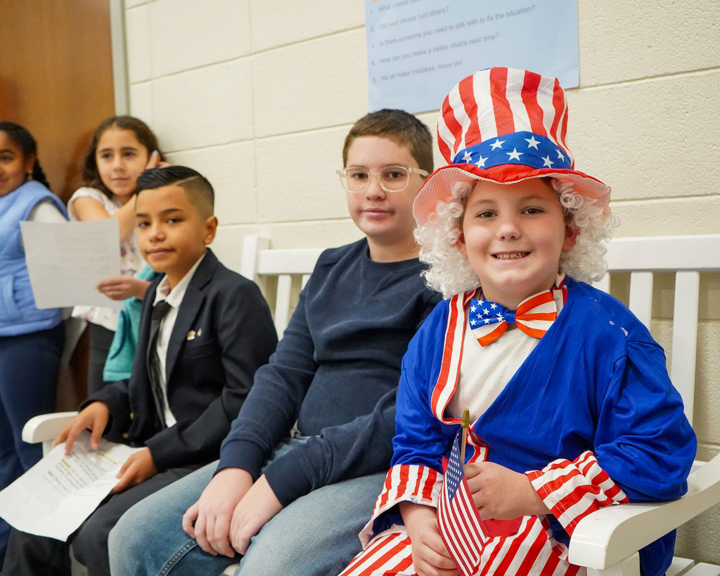 Students are sitting on a bench at a Veterans Day Program. One Student is dressed up in patriotic gear and smiling at the camera. 