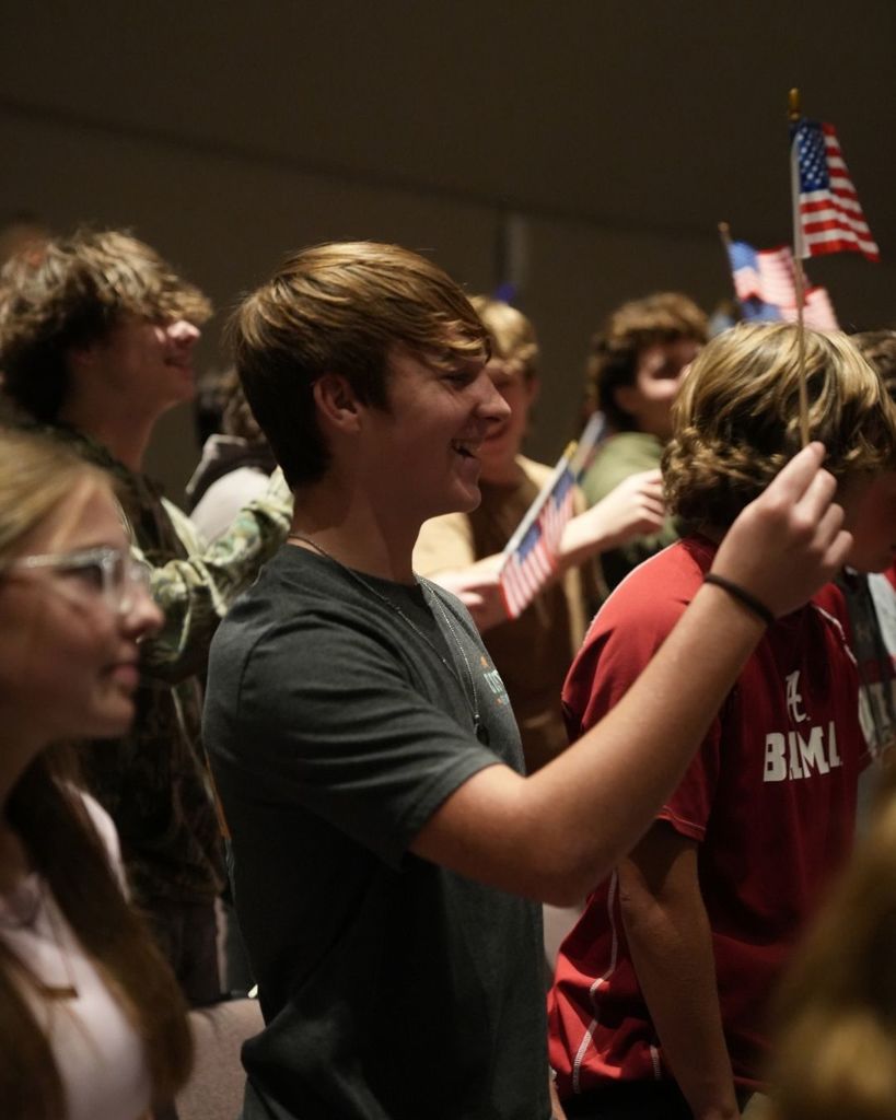 Students wave small American flags while standing in a school auditorium.