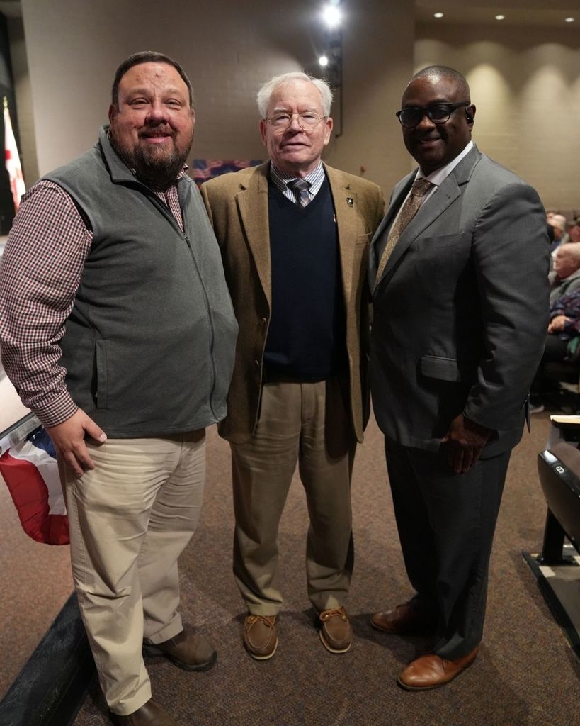 Three men stand together and pose for a photo inside a school auditorium.