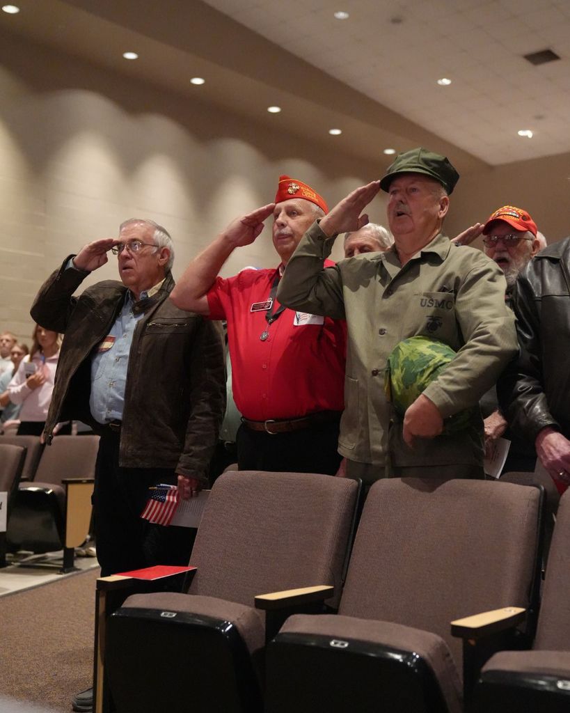 Veterans salute while standing in a school auditorium.