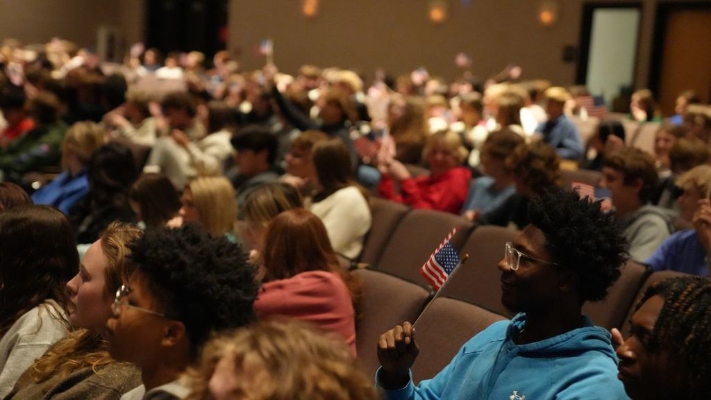 Students hold small American Flags while sitting in a school auditorium.