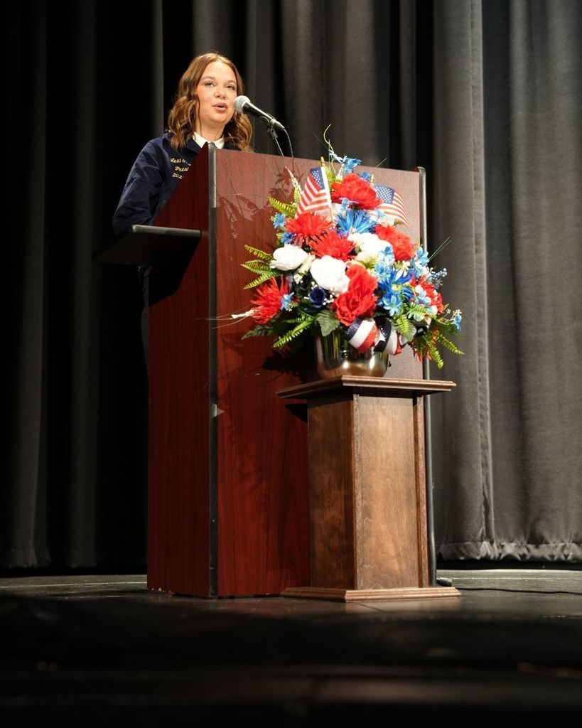 A student wearing an FFA Jacket speaks into a microphone on a stage inside a school auditorium.