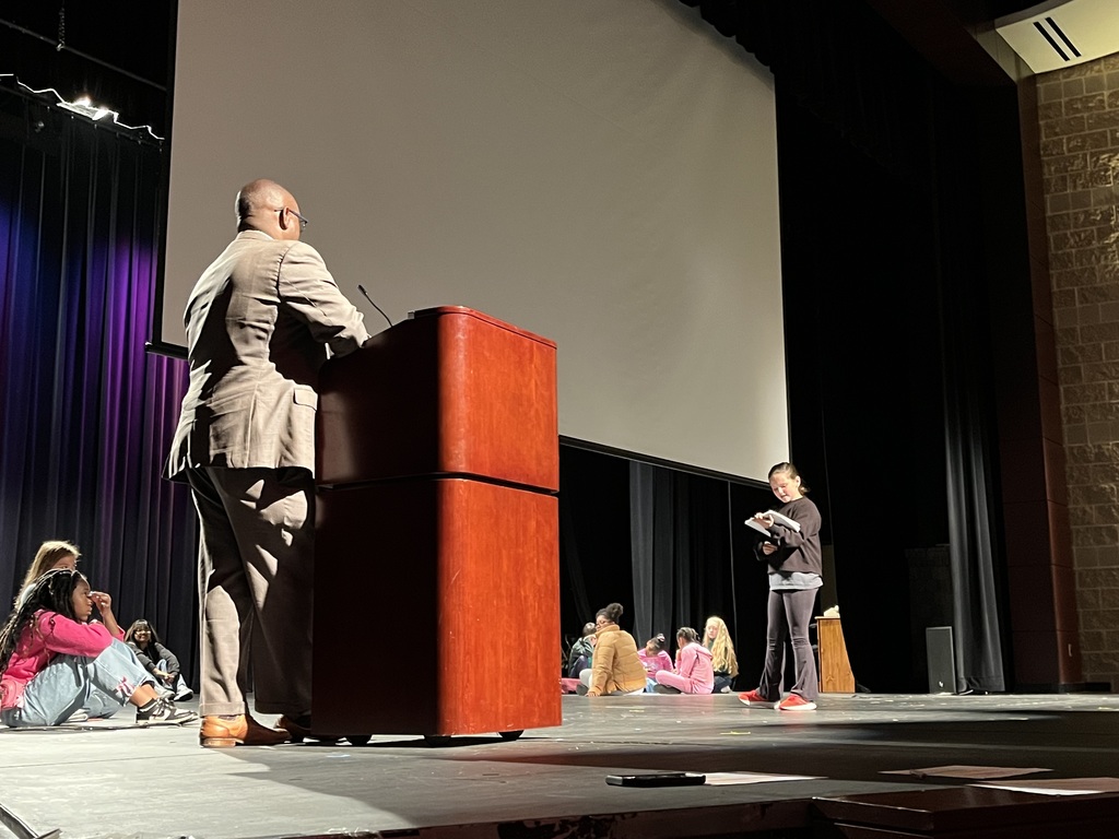 JEFCOED Superintendent Dr. Gonsoulin stands at a podium on a stage listening to a student speak during the Superintendent's Student Advisory Council Meeting. Other students sit on the stage and listen to the student speak as well.