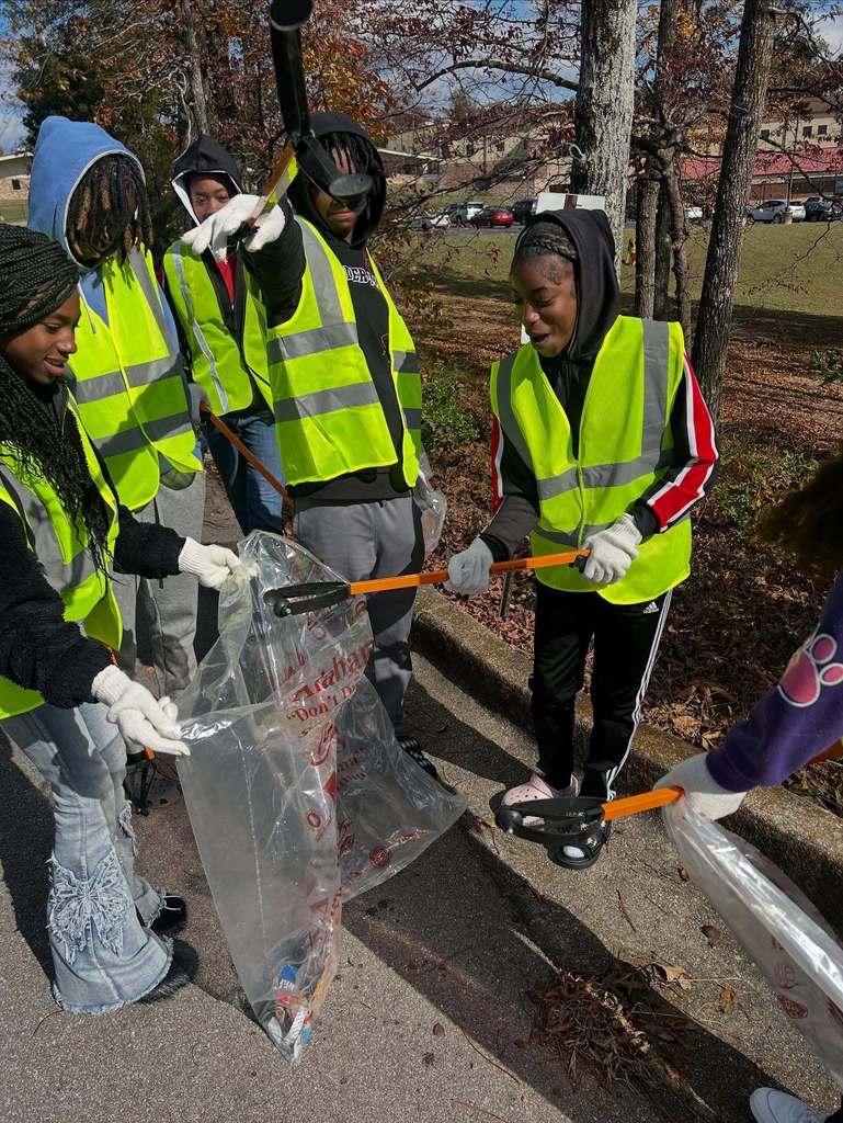 Students wearing yellow vests put trash into a trash bag while standing outside a school.