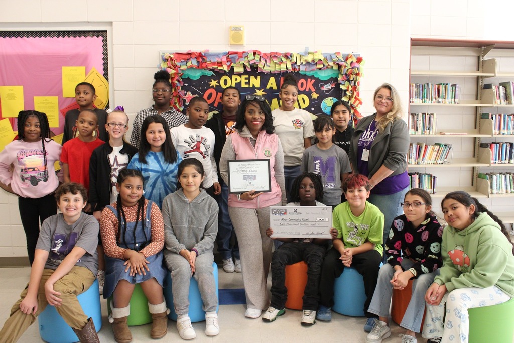 A group of students and two adults pose for a photo inside a library. The adult in the middle, Dr. Kimberly Hudson Smith holds an award. A child next to her holds a check for a grant.