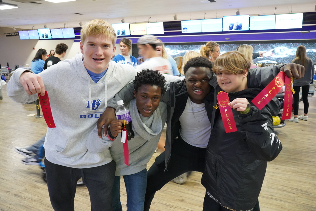 Four students stand together for a photo at a bowling alley. They smile and hold up red ribbons.
