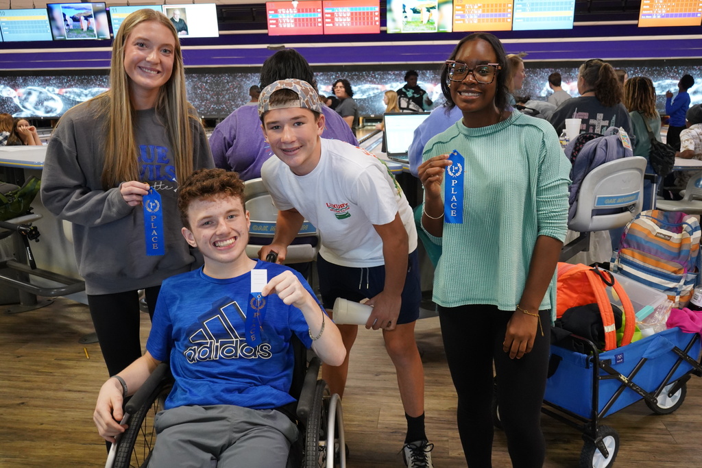 Students smile and hold up blue ribbons while standing and sitting inside a bowling alley.