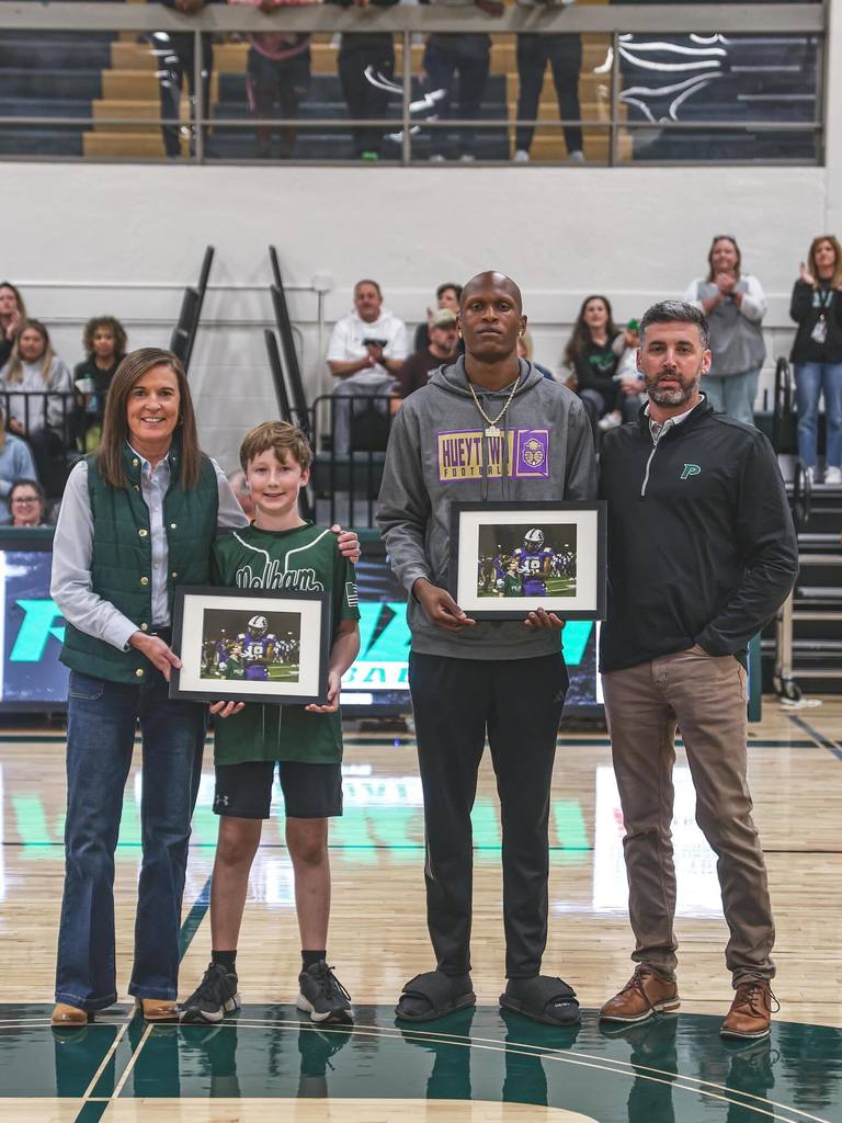 Two adults and two students pose for a photo in a school gym. The students hold framed photos in the picture.