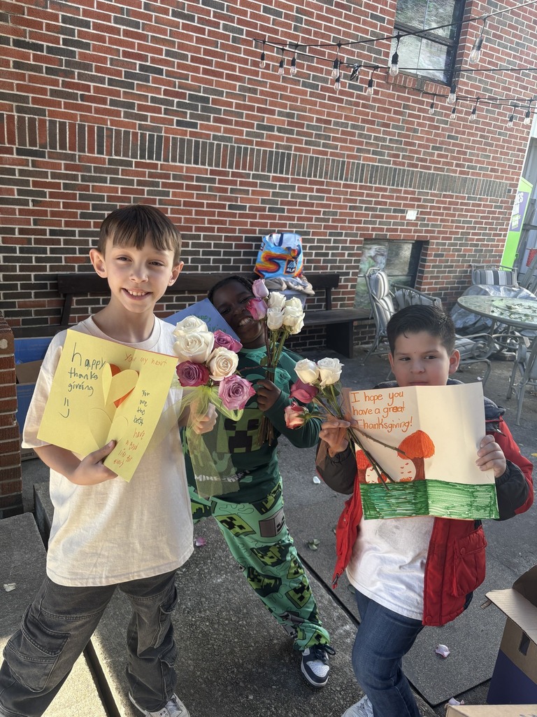 Students smile and hold up hand-drawn cards while standing outside a brick building.