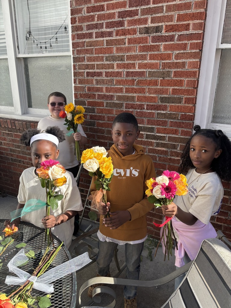 Students stand outside a brick building and hold up bouqets of flowers.