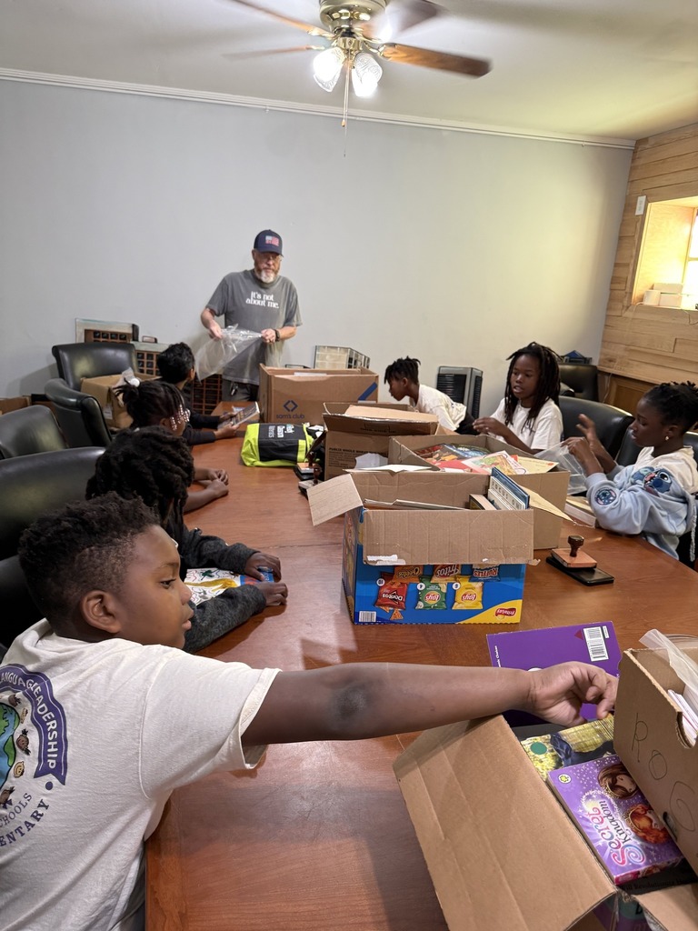 Students sit at a table inside a room and pack supplies into boxes.