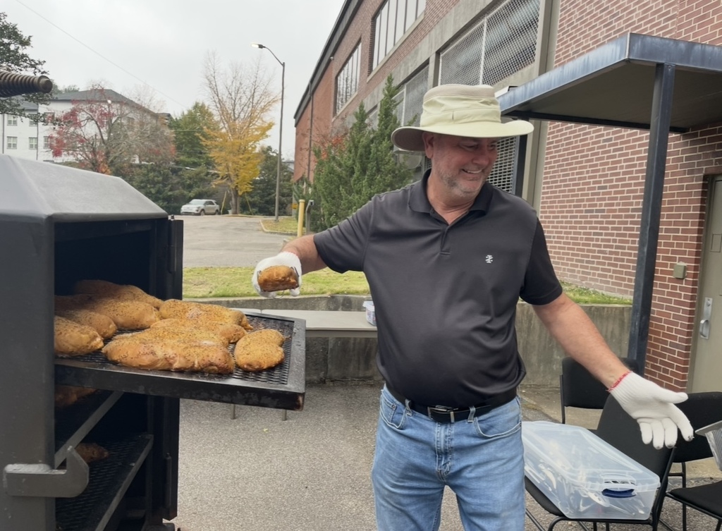 A man smiles and uses tongs to pull a chicken out of a BBQ smoker. The man is standing in a parking lot outside the Jefferson County Board of Education.