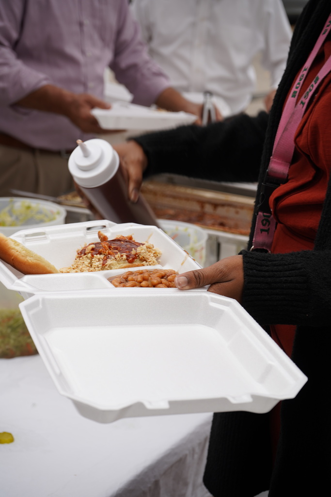The photo is a close up of a woman holding a to-go container with a barbecue sandwich and baked beans on it.