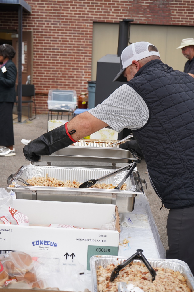 A man wears oven mits and holds a large container while standing at a table filled with containers of BBQ.