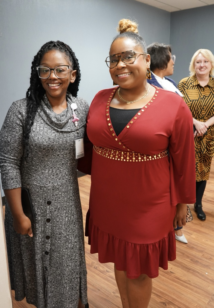 Two women stand together and smile for a photo while standing inside a hallway.