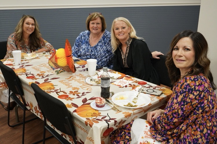 Four women sit together at a table and smile while eating a Thanksgiving lunch.