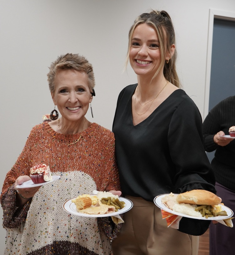 Two women stand together for a photo and smile while holding plates with Thanksgiving food on them.