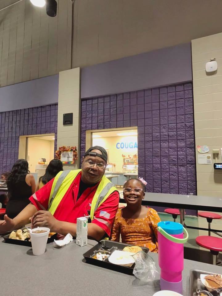 An adult and a child sit together at a lunch room table and smile for a photo.