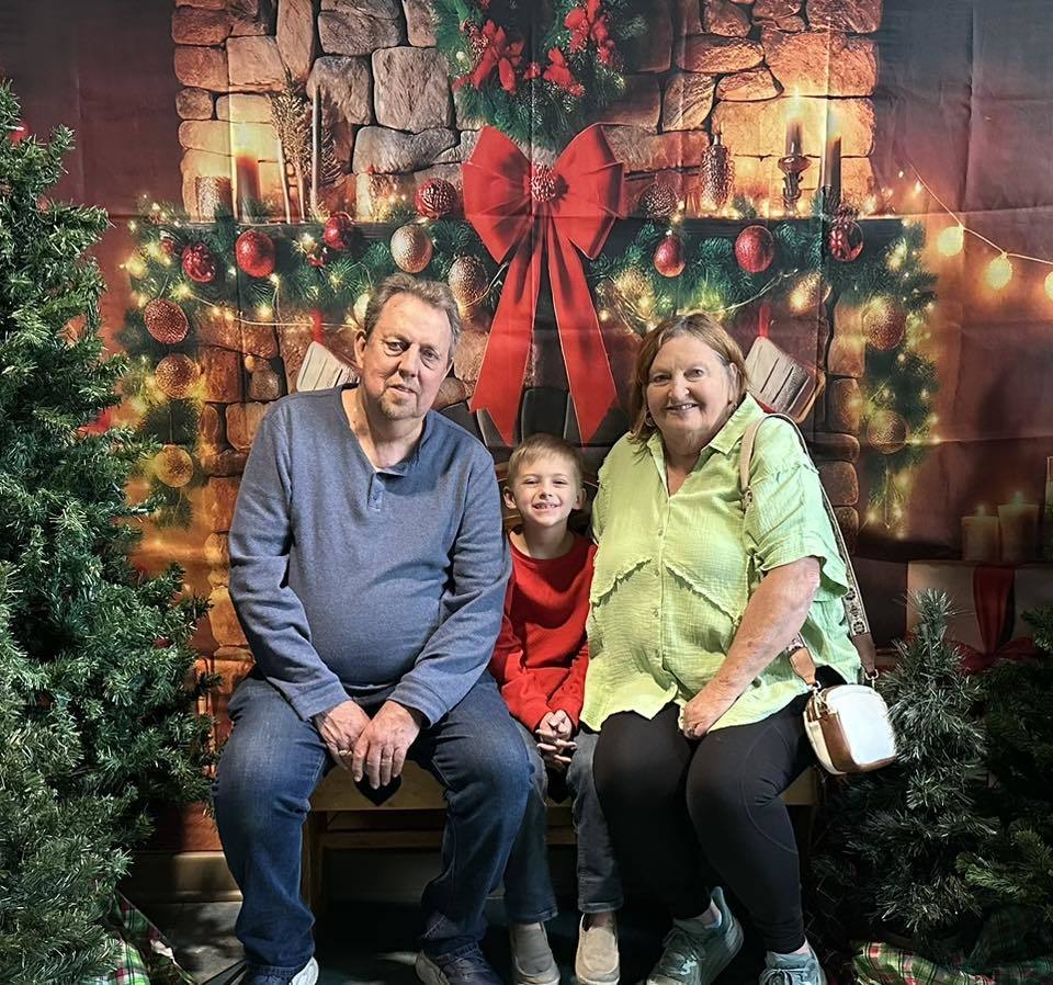 Two adults and a child sit together for a photo in front of a Christmas backdrop. 