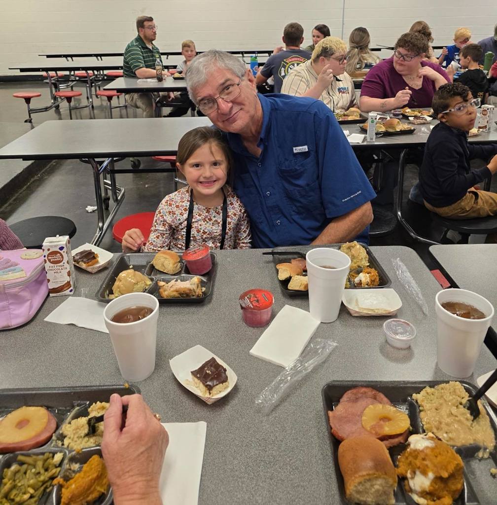 An adult and a child sit together at a school lunchroom table and smile for a photo.