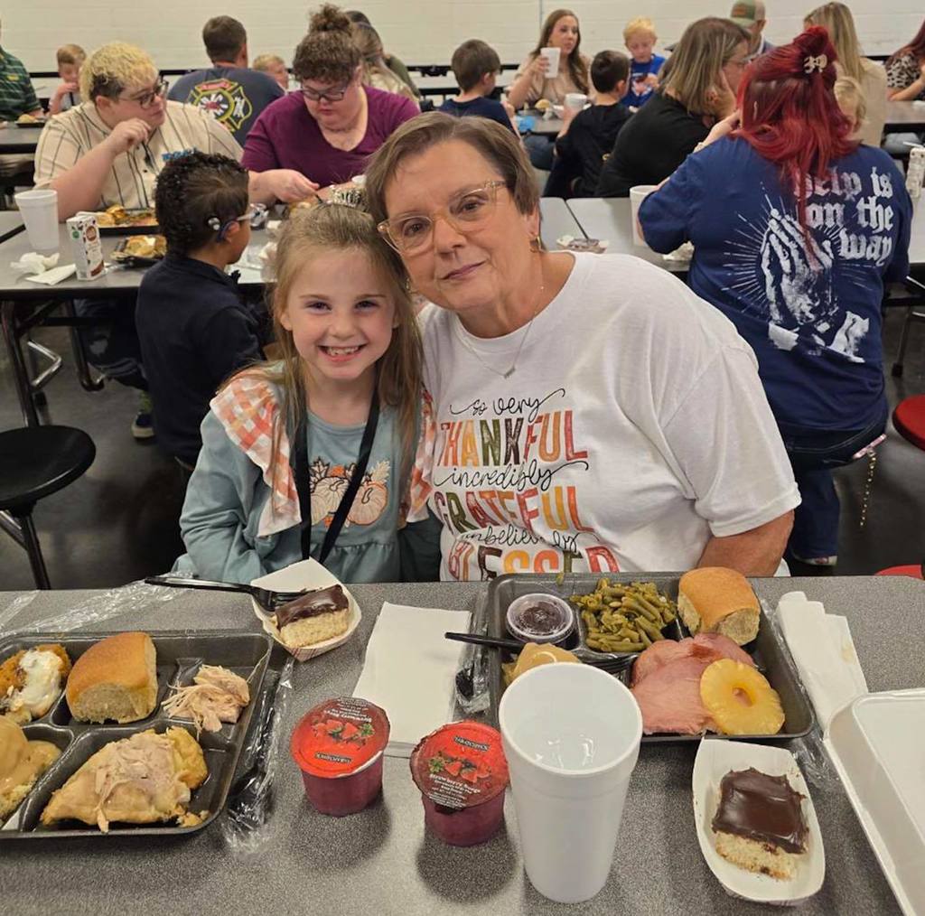 An adult and a child sit together at a school lunchroom table and smile for a photo.
