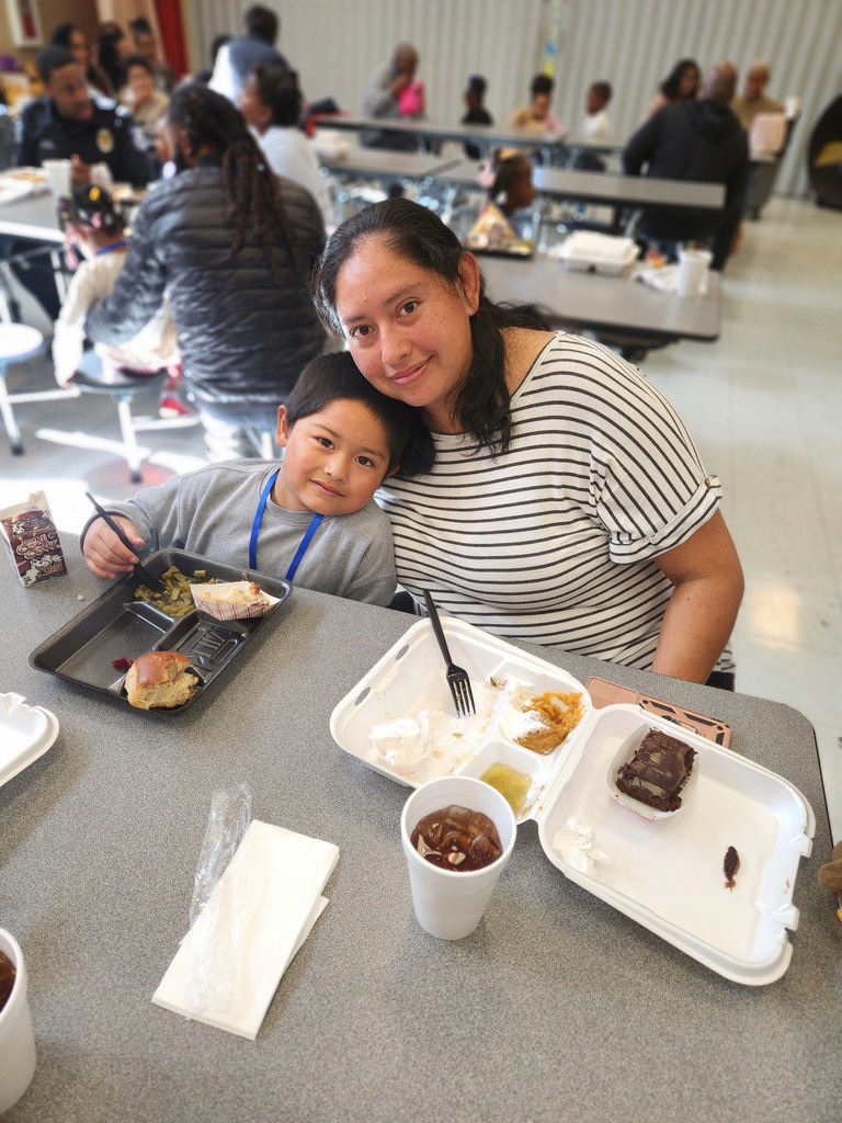 An adult and a child sit together at a school lunch table and smile for a photo.