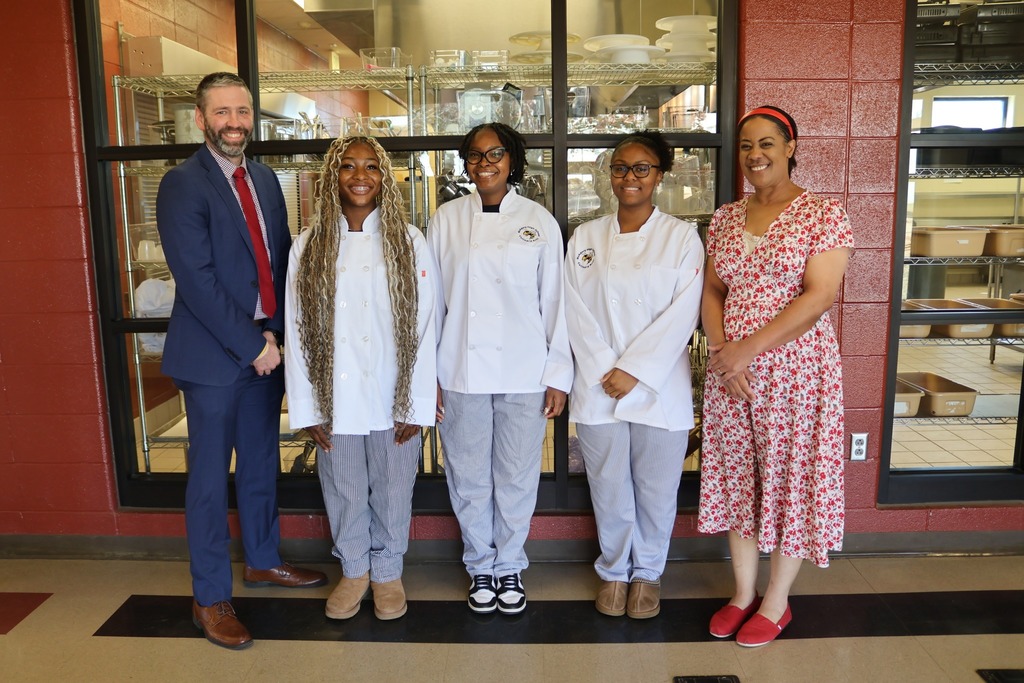 Two adults and three culinary students wearing white coats stand togther for a photo and smile.