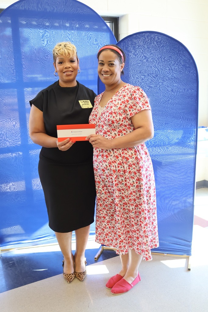 Two women stand together in a school hallway and smile while holding a check.