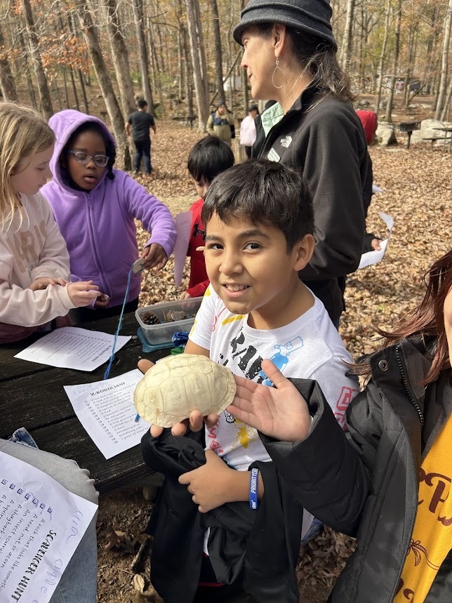 Students and an adult stand around a table outside with pieces of paper on it. One of the students holds up a turtle shell.